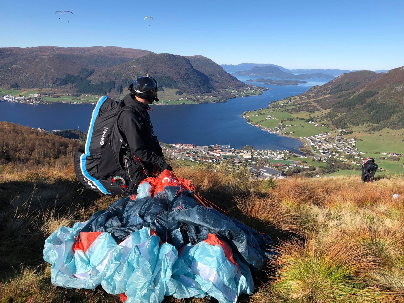 A paraglider pilot preparing equipment at a mountain launch above a fjord, with other paragliders in the air.