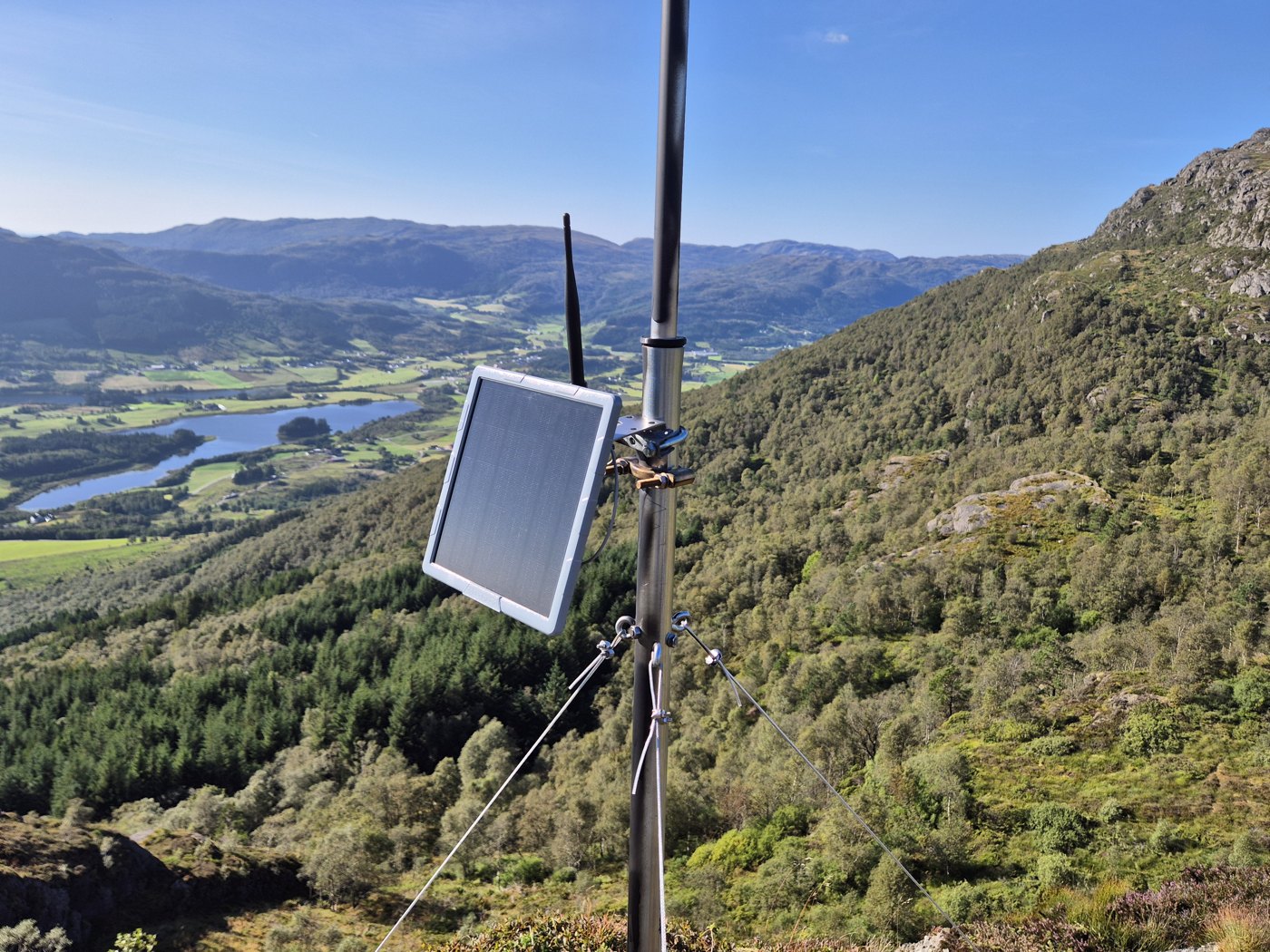 Solar node with panel and radio antenna mounted on a mast outdoors.